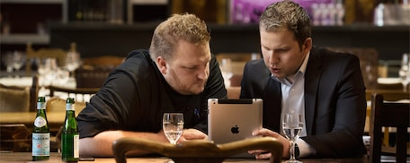 Zwei Männer sitzen am Tisch und arbeiten gemeinsam mit ihrem iPad an ihrem Businessplan für ihre Gastronomie. 
