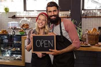 Zwei Gastronomen, die ein „Geöffnet“-Schild hochalten –aktuelle Informationen sollten auch auf der Website der eigenen Gastronomie prominent abgebildet werden.