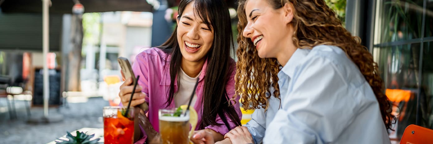 Deux jeunes femmes riant en regardant un smartphone à la terrasse d’un café