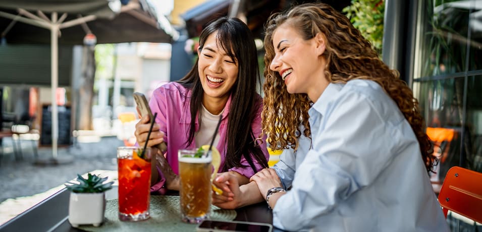 Deux jeunes femmes riant en regardant un smartphone à la terrasse d’un café