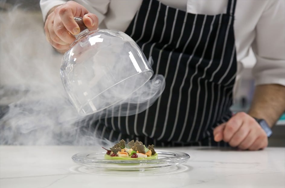Chef soulève une cloche en verre remplie de fumée au dessus d’un plat