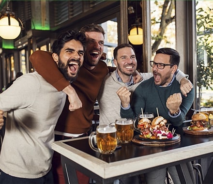 Groupe d'amis dans un restaurant, avec des burgers et une bière devant une retransmission sportive