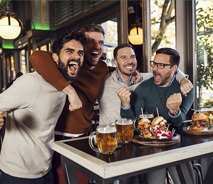Groupe d'amis dans un restaurant, mangeant des burgers et buvant une bière devant une retransmission sportive