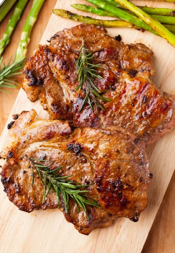 Două bucăți de ceafă de porc gătită la grătar, rumenite și suculente, decorate cu crenguțe de rozmarin, așezate pe un tocător din lemn; în fundal se văd fire de sparanghel verde