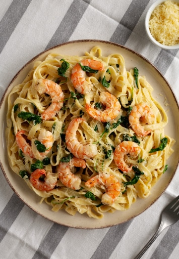 Farfurie cu tagliatelle cremoase, creveți rumeniți și frunze de spanac, servită cu parmezan ras, pe șervet alb cu dungi gri.