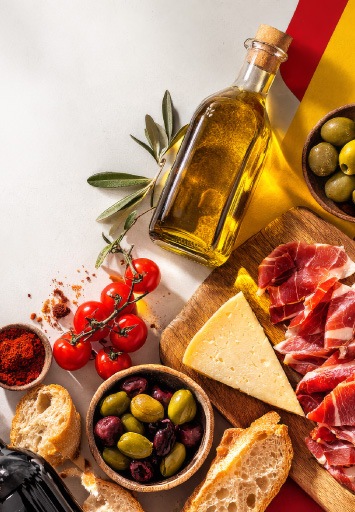 Platou cu specialități spaniole: ulei de măsline, măsline, jamón, brânză maturată și pâine artizanală, pe fundal inspirat de steagul Spaniei.