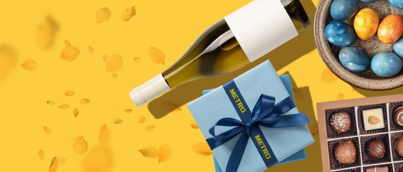 Set cadou de Paște METRO cu sticlă de vin, cutie albastră și ouă colorate, expuse elegant pe fundal galben.