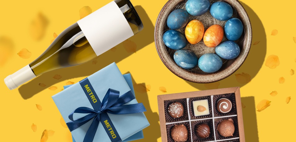 Colecție METRO de cadouri pentru Paște cu vin alb, cutie albastră, ouă încondeiate și praline, pe fundal galben.