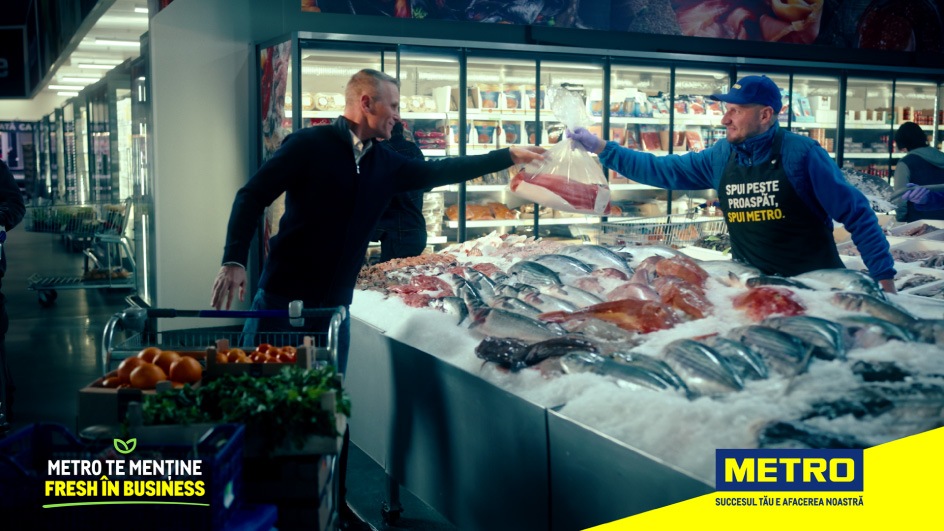 Scene din raionul de pește proaspăt din magazinul METRO: un client primește un produs direct de la un angajat, în fața unui tejghea plină cu pește și fructe de mare, sub mesajul «METRO – Succesul tău e afacerea noastră».