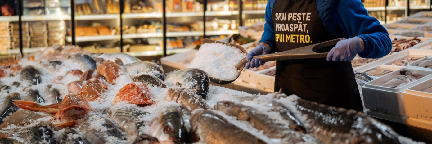 Raionul de pește proaspăt din METRO, unde un angajat adaugă gheață pentru menținerea calității produselor. Sortiment variat de pește și fructe de mare, prezentat pe pat de gheață pentru prospețime optimă.