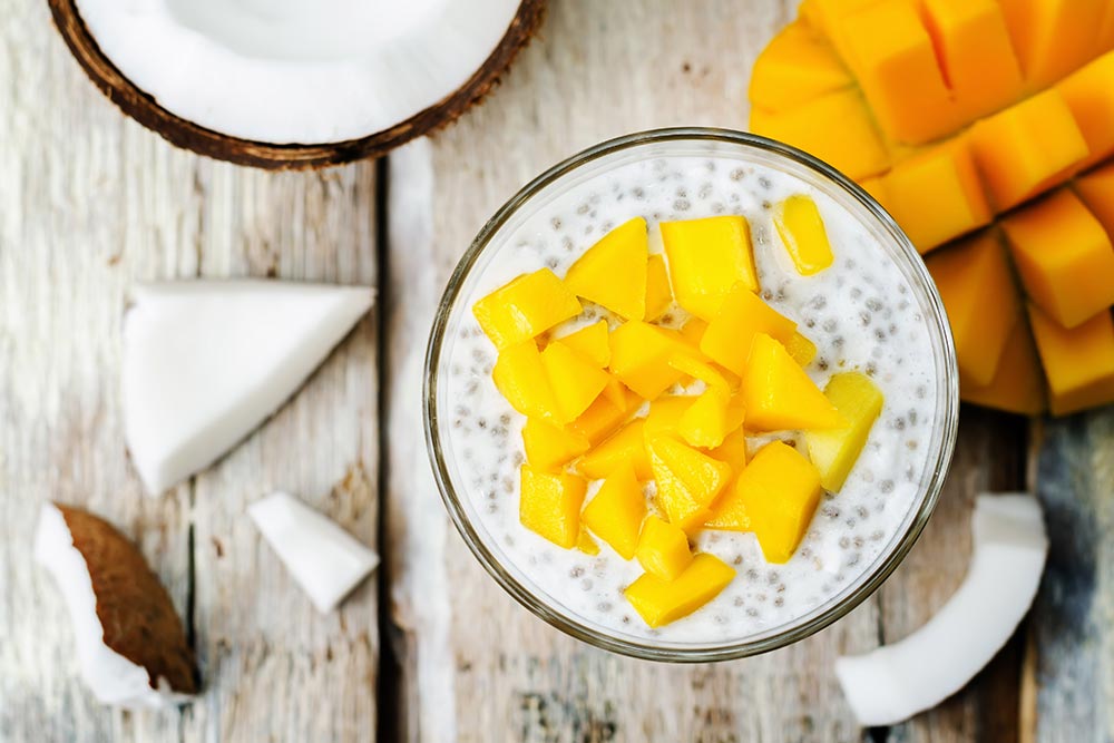 Чіа-пудинг із шматочками свіжого манго в склянці, оточеній кокосом і манго.