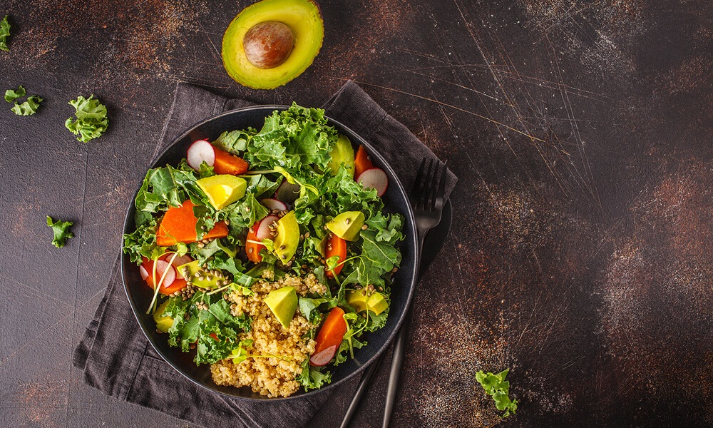 Салат з капустою кале, авокадо, помідорами, редисом і кіноа – яскравий овочевий салат у чорній тарілці на темному фоні.