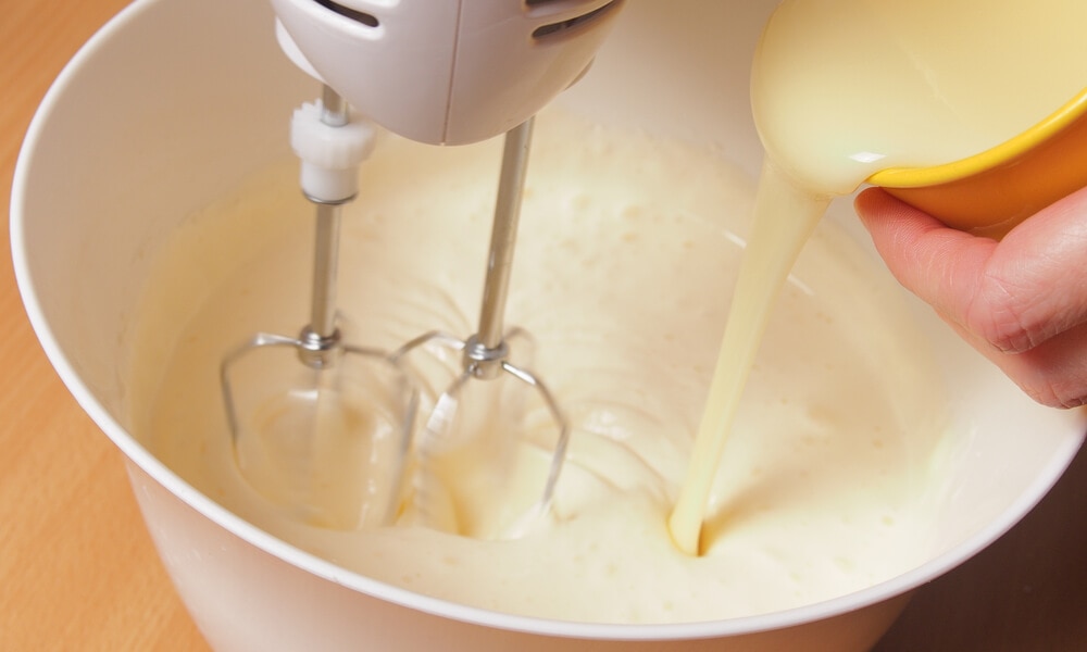 Процес приготування крему: у збиті вершки в чаші міксера додається згущене молоко з жовтої чашки