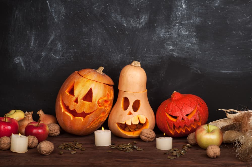 Три вирізані гарбузи з підсвіткою, свічки, яблука та горіхи на фоні шкільної дошки.