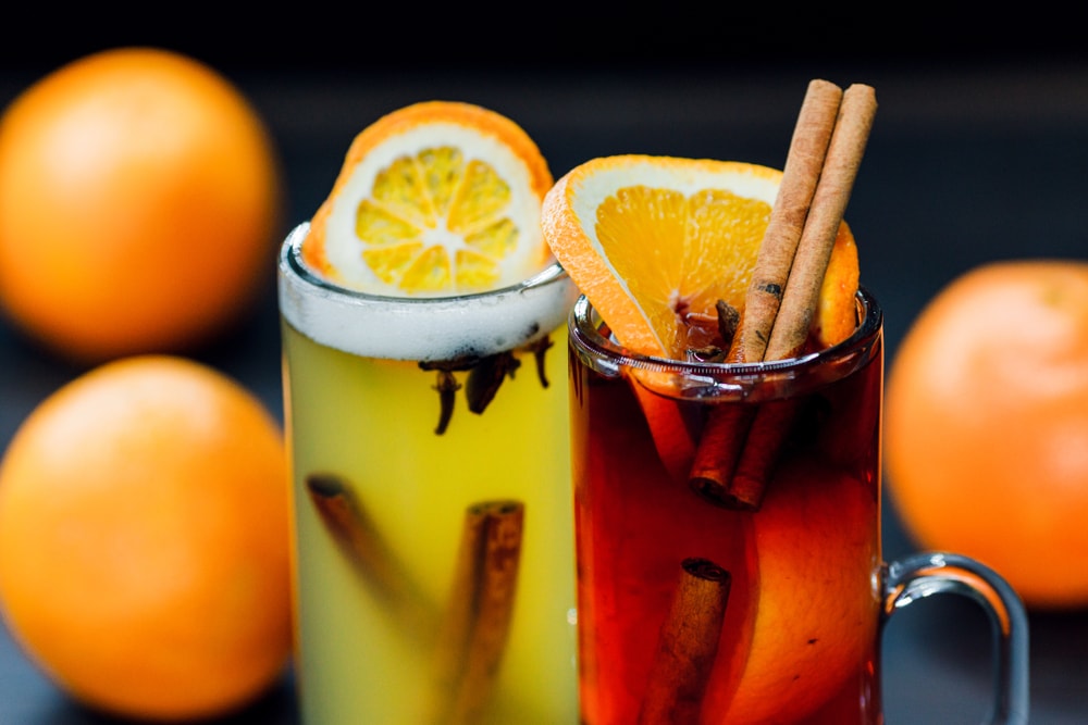 Два різнокольорові глінтвейни з апельсином, корицею та гвоздикою на фоні апельсинів