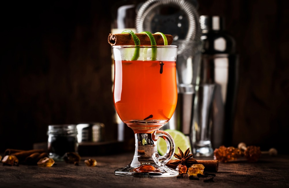 Гарячий коктейль із корицею та лаймом у прозорій келиховій склянці на фоні барного інвентарю
