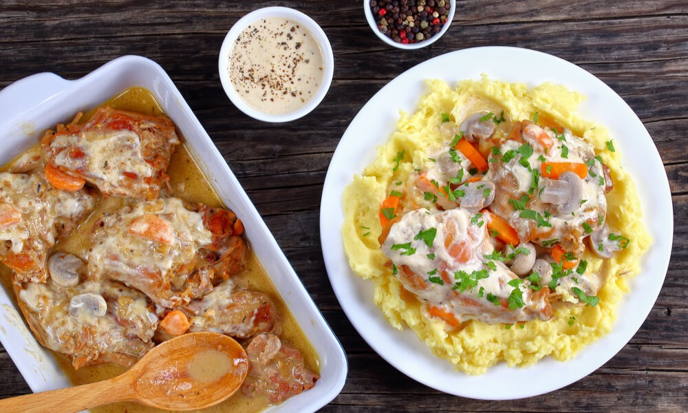 Курка, тушкована з овочами та грибами в вершковому соусі, подана з картопляним пюре на тарілці, поруч форма з готовою стравою та соусом