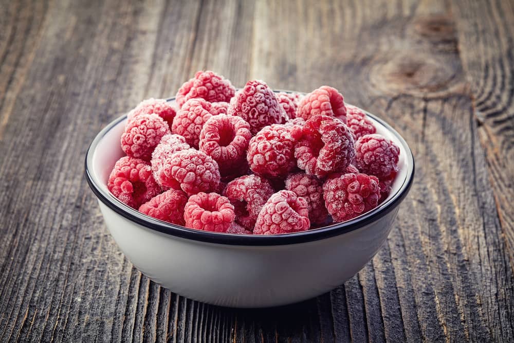 біла керамічна миска, наповнена замороженою малиною на дерев’яному столі