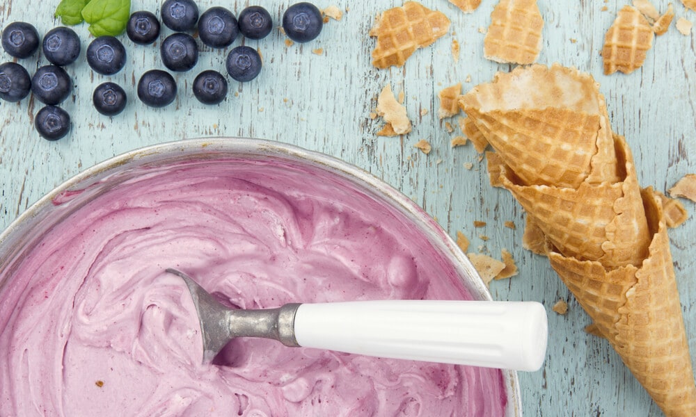Миска з ягідним морозивом і ложкою, поруч вафельні ріжки та свіжі чорниці на дерев'яному столі