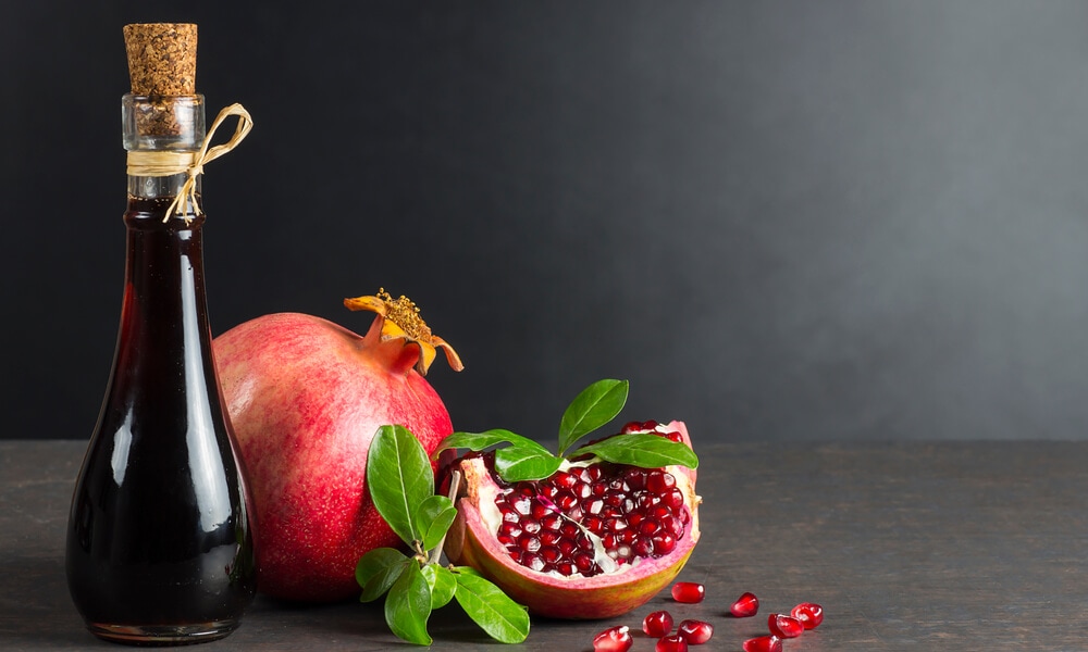 Пляшка з гранатовим соусом поряд із цілим та розрізаним гранатом на темному фоні.