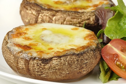 Великі печериці, запечені з сиром, подані з листям салату та половинкою томата на білому фоні