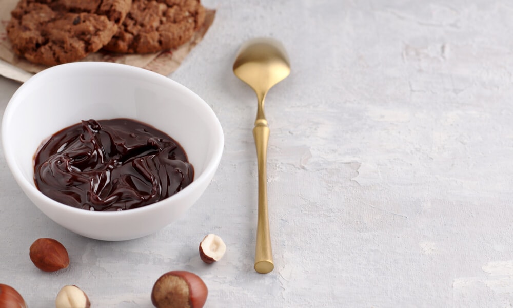 Шоколадна паста в білому посуді поруч із печивом і ліщиною