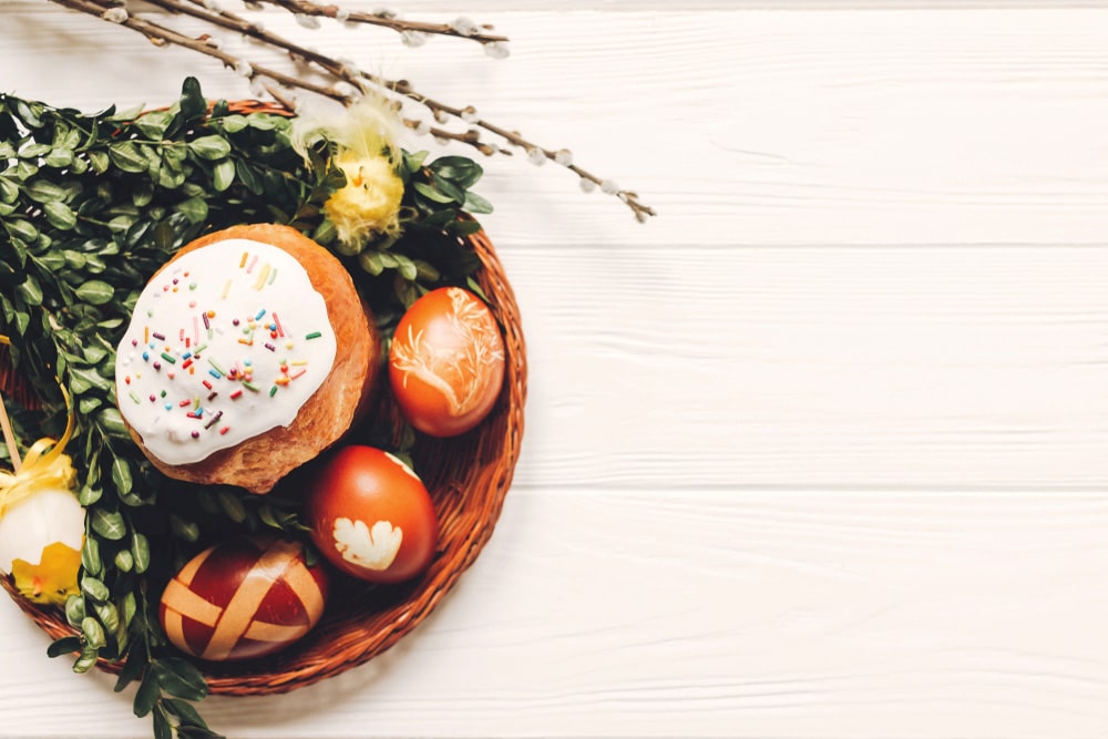 Великодній кошик з паскою та розписаними яйцями на білому дерев’яному столі.