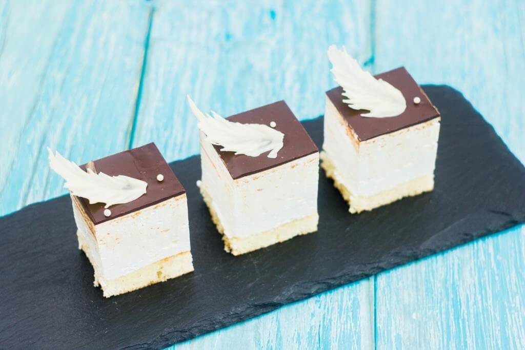 Три міні-десерти з білим кремом і шоколадною глазур'ю, прикрашені білими шоколадними пір'їнами, на чорній сланцевій підставці.