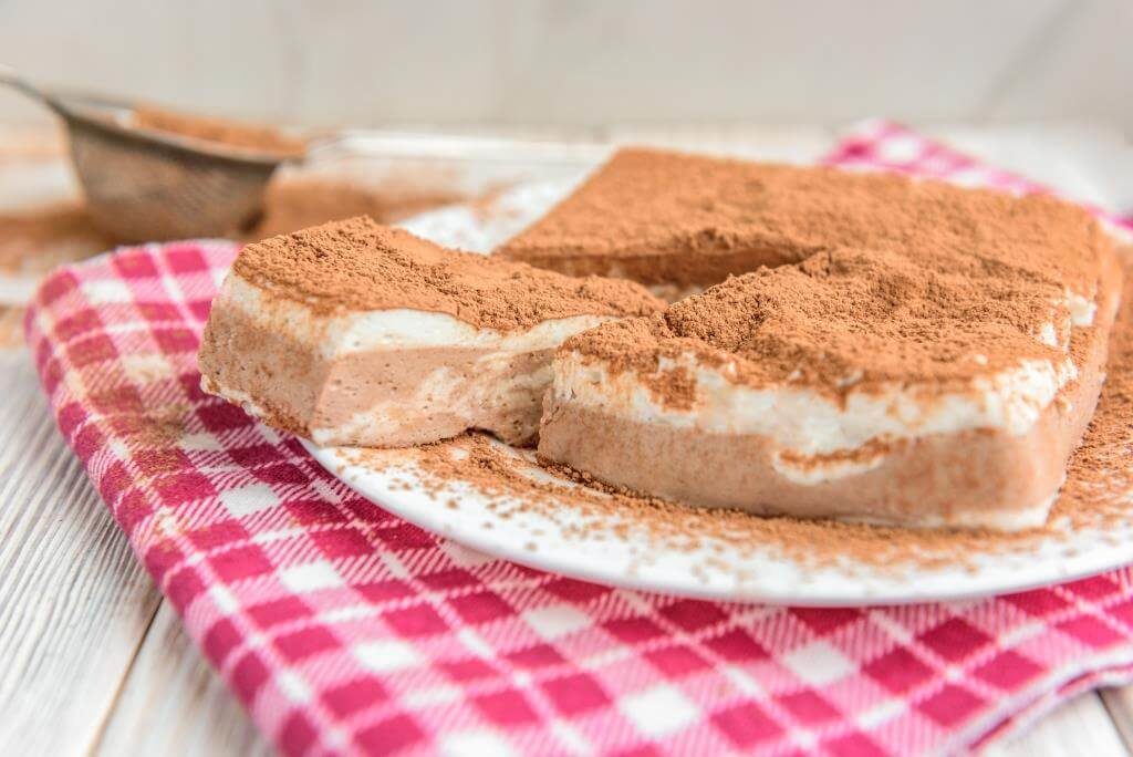 Десерт з какао і вершковим кремом на тарілці, прикрашений порошком какао, на рожевій серветці в клітинку.