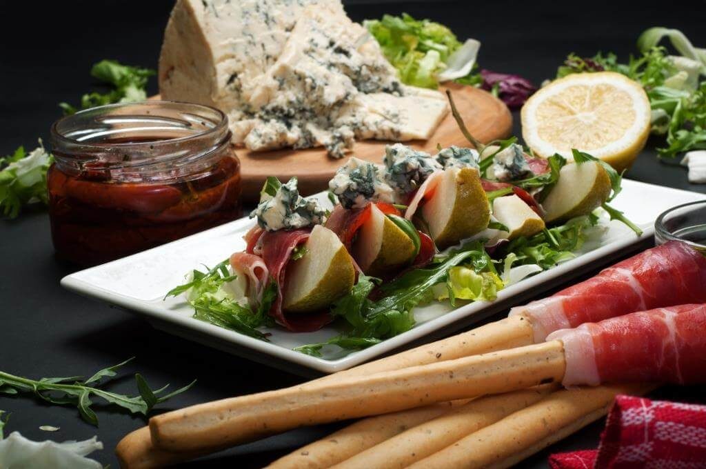 Закуска з груші, прошуто та блакитного сиру на листі салату, поруч грісіні та баночка в'ялених томатів