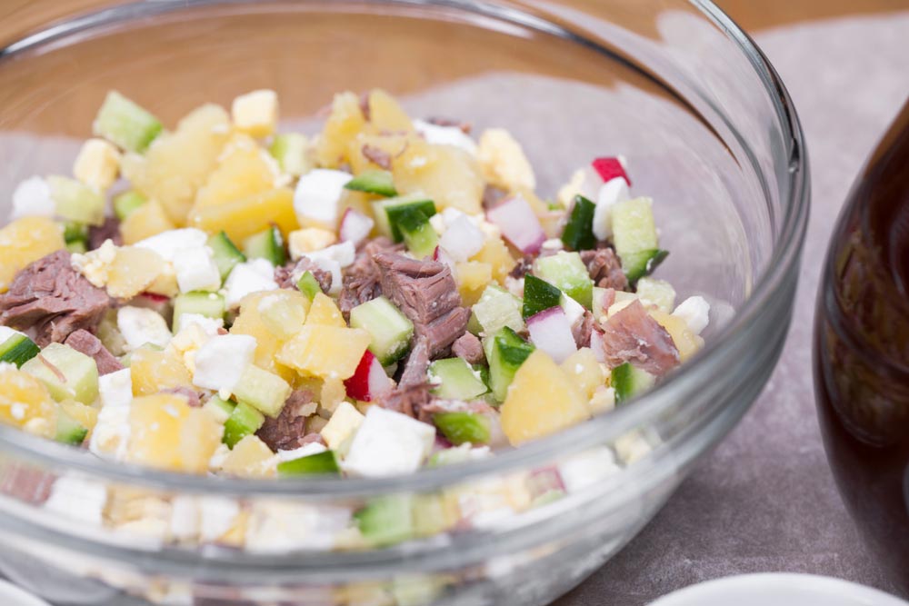 Нарізані інгредієнти для окрошки: картопля, огірки, редис, яйця, яловичина та зелень у скляній мисці