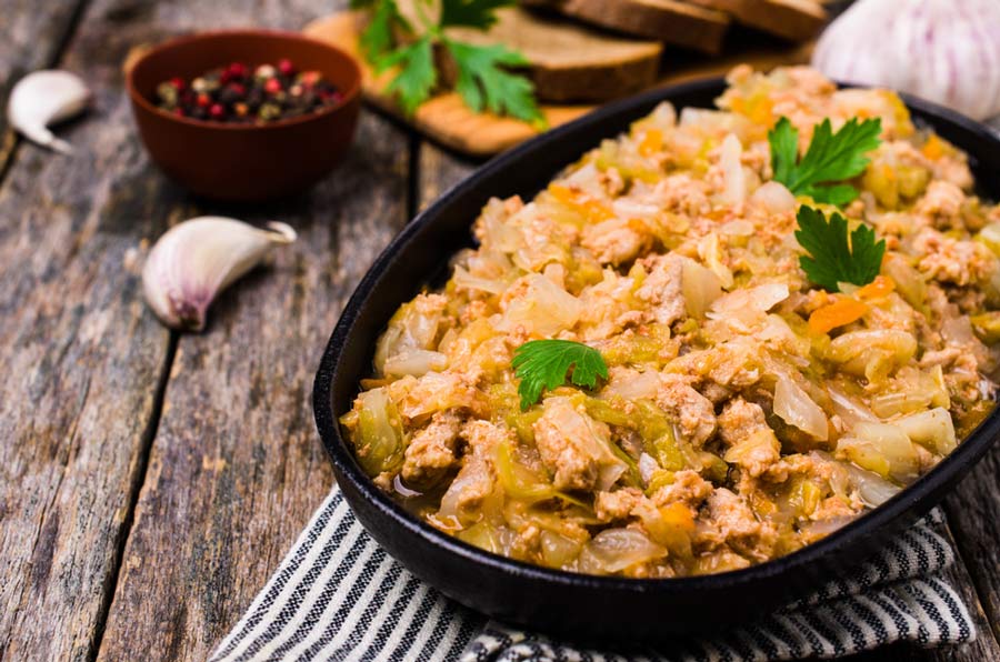 Тушкована капуста з м’ясом у чорній мисці на дерев’яному столі, поруч часник і спеції.