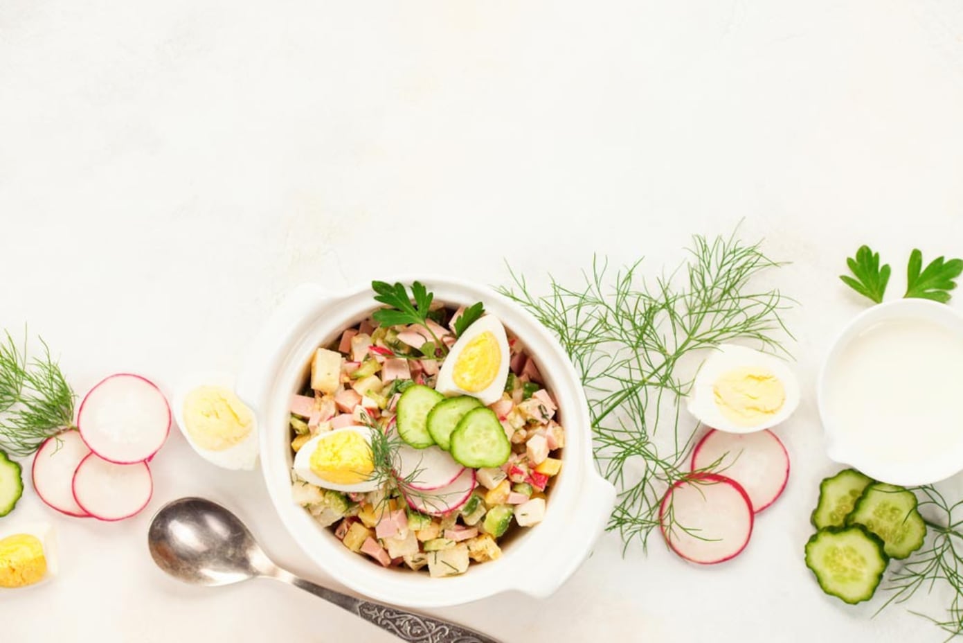 Салат окрошка з ковбасою, огірками, редискою та яйцями в глибокій білій тарілці на світлому фоні.