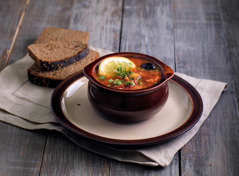 Глиняний горщик із солянкою, лимоном та оливкою на тарілці, поруч скибки житнього хліба