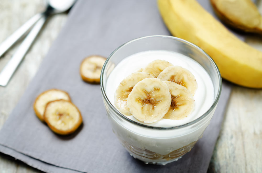 Десерт із йогуртом і бананом у склянці, на фоні банан та сервірувальні ложки