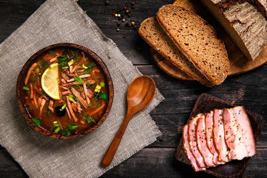 Тарілка м'ясної солянки з лимоном і зеленню, поруч чорний хліб і нарізане сало