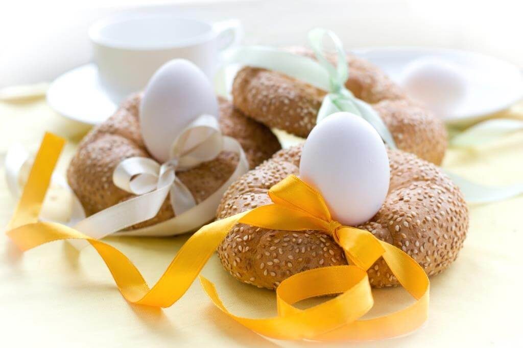 Пасхальні булочки з кунжутом у формі вінків із білими яйцями всередині, прикрашені кольоровими стрічками