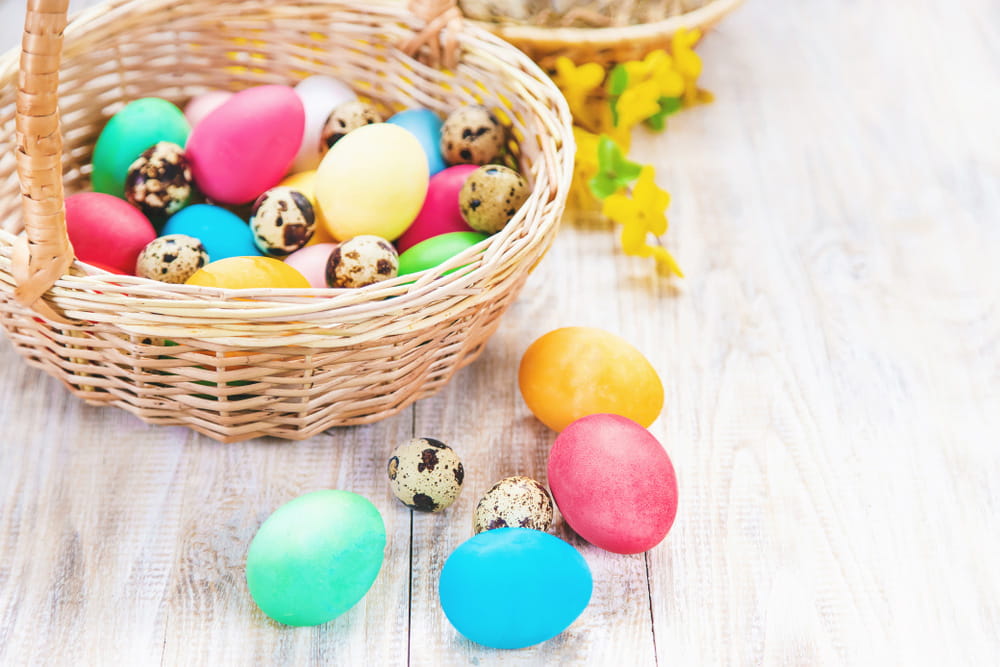 Кошик з кольоровими великодніми яйцями та перепелиними яйцями на світлому дерев’яному столі.
