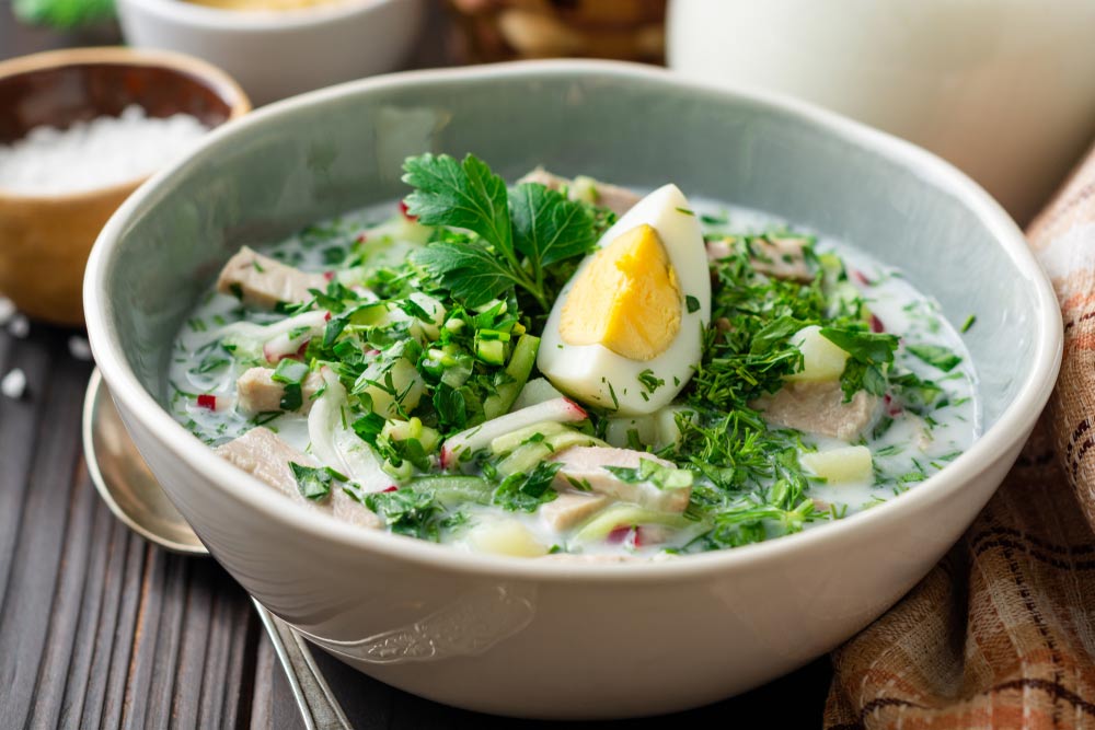 Класична окрошка на кефірі з вареним яйцем, зеленню та ковбасою в керамічній мисці.
