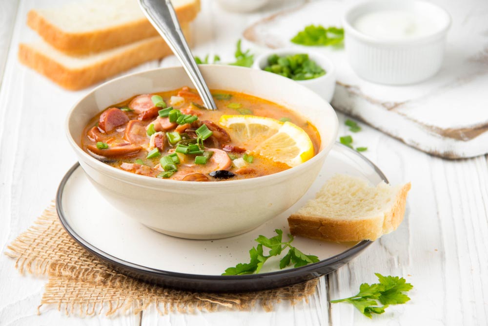 Світла миска з солянкою, шматочком лимона та зеленню, подана з білим хлібом