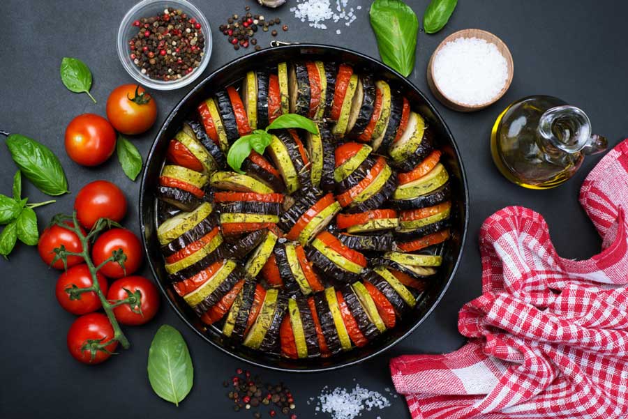Класичний рататуй з баклажанами, помідорами та цукіні, красиво викладений у круглій чорній формі, поруч із помідорами, базиліком, олією та спеціями.
