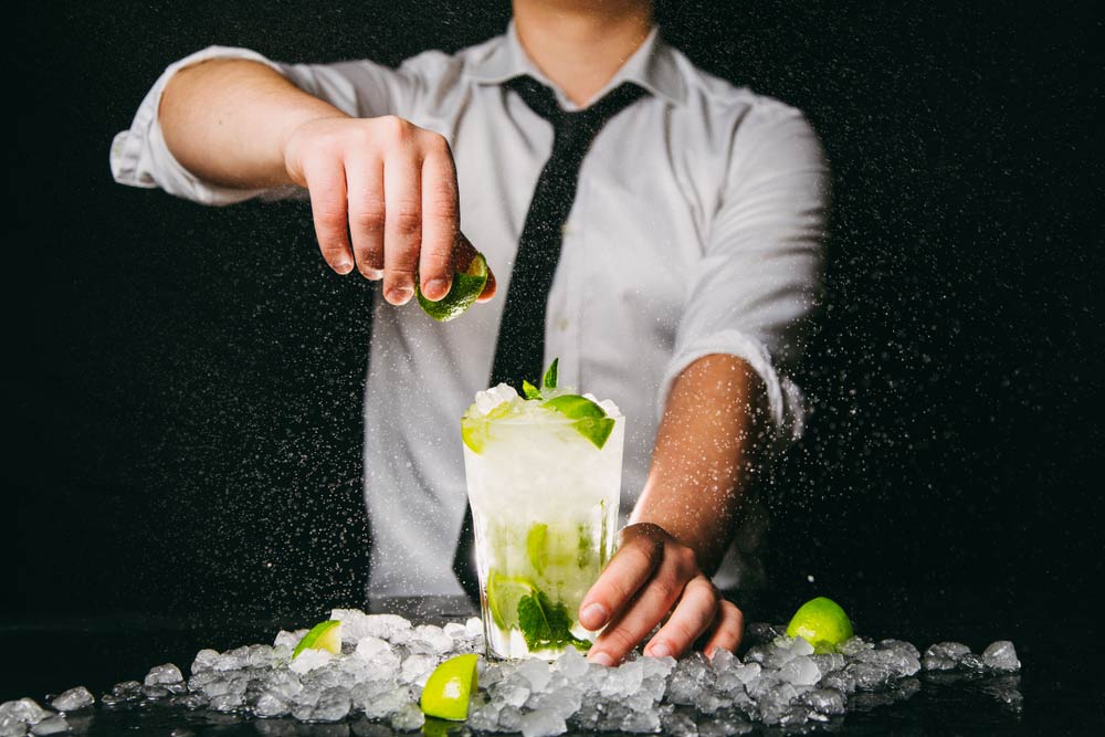 Бармен посипає вологу склянку з мохіто льодом, стискаючи лайм, фон — чорний.