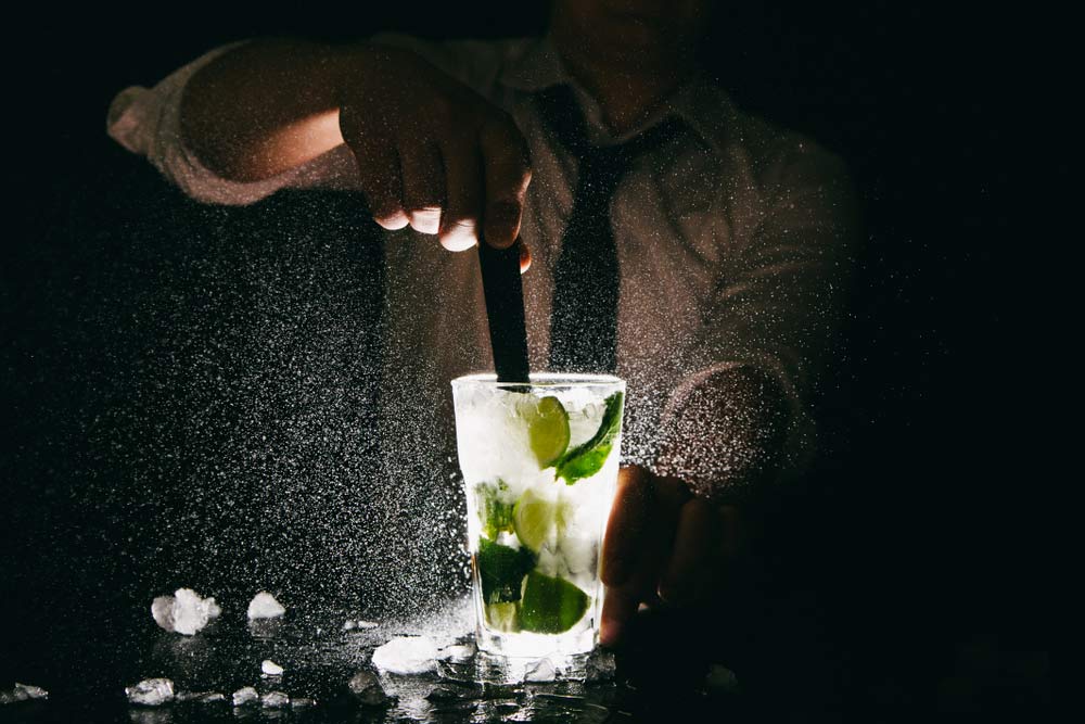 Бармен перемішує коктейль мохіто, розлітаються краплі води й льоду на фоні темряви.