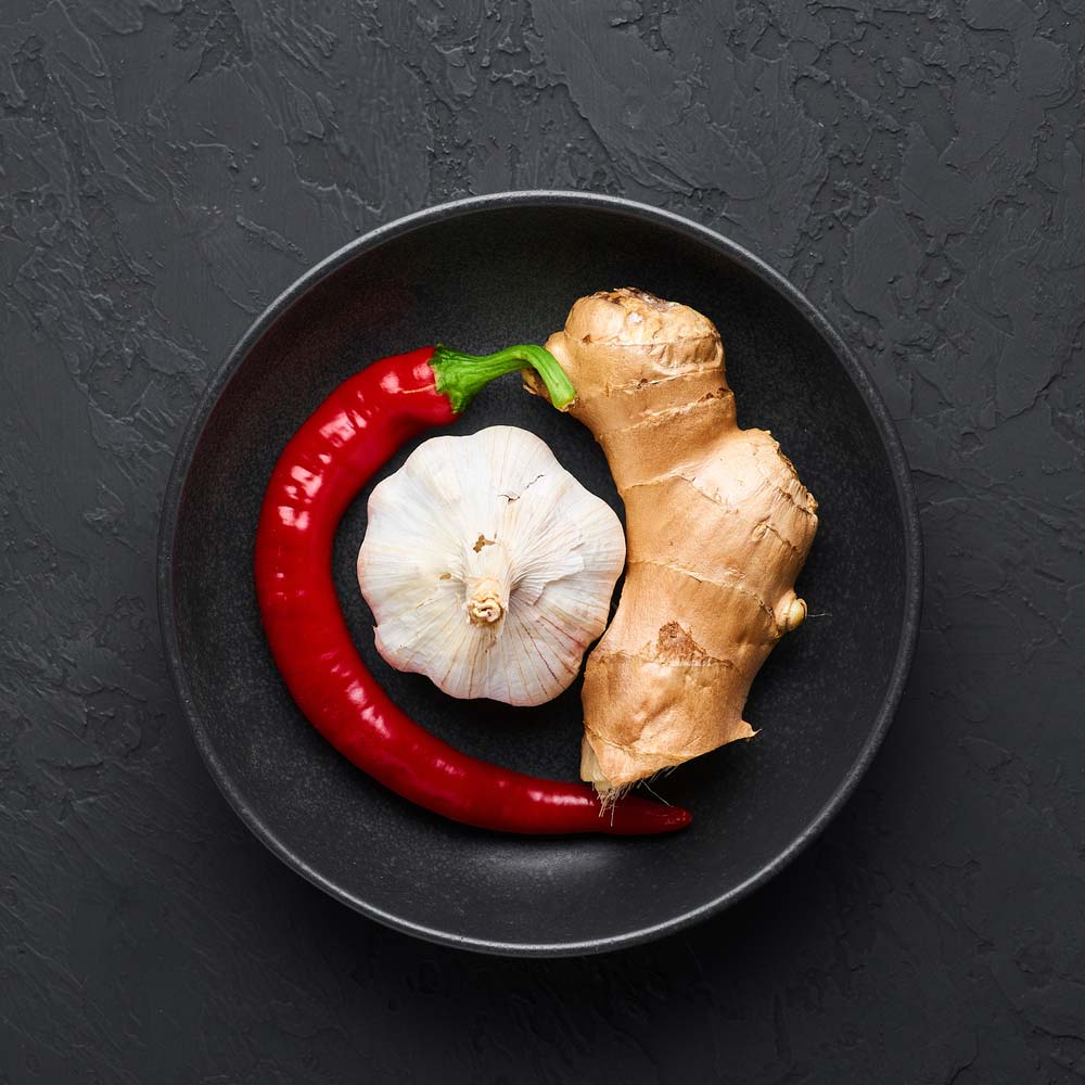 Імбир, головка часнику та стручок червоного перцю в чорній тарілці на сірому фоні