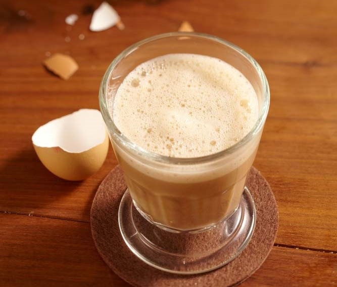 Склянка з традиційним яйцево-кавовим напоєм на дерев'яному столі, поруч лежать уламки яєчної шкарлупи.