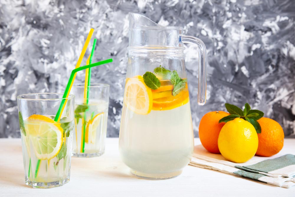 Глечик і склянки з лимонадом, лимоном, апельсином і м'ятою на білому фоні.