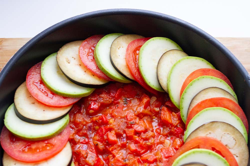 Скибочки баклажанів, кабачків і помідорів викладені по колу на соусі в чорній формі для запікання, підготовка до приготування рататуя.