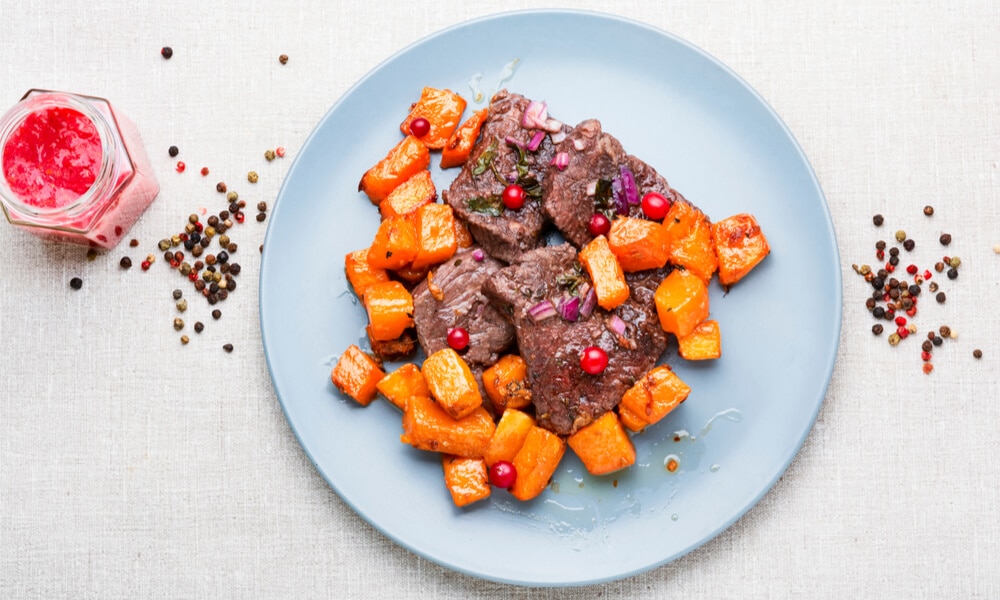 Тушкована яловичина з гарбузом та брусницею на блакитній тарілці, поруч розсипані зерна перцю та соус у баночці.