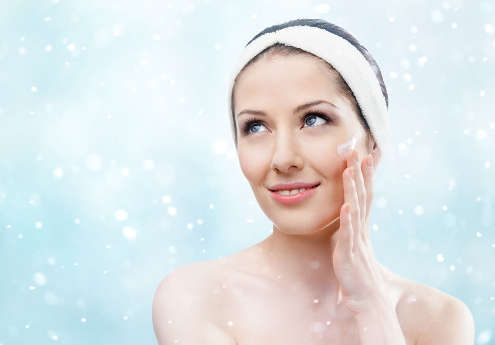 Жінка з білою пов'язкою на голові наносить крем на обличчя на фоні сніжинок.