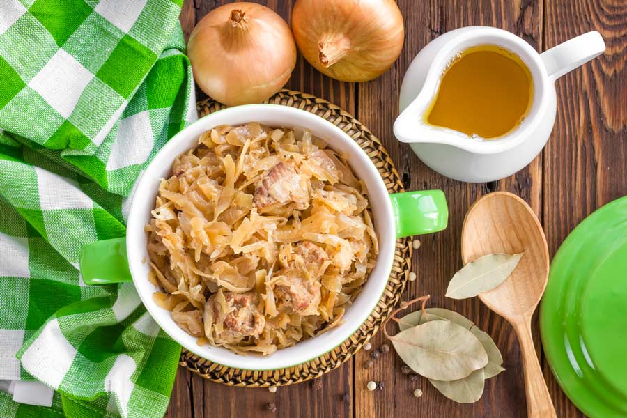 Капуста з м’ясом у зеленій каструлі, на столі з цибулею, лавровим листом і дерев’яною ложкою.